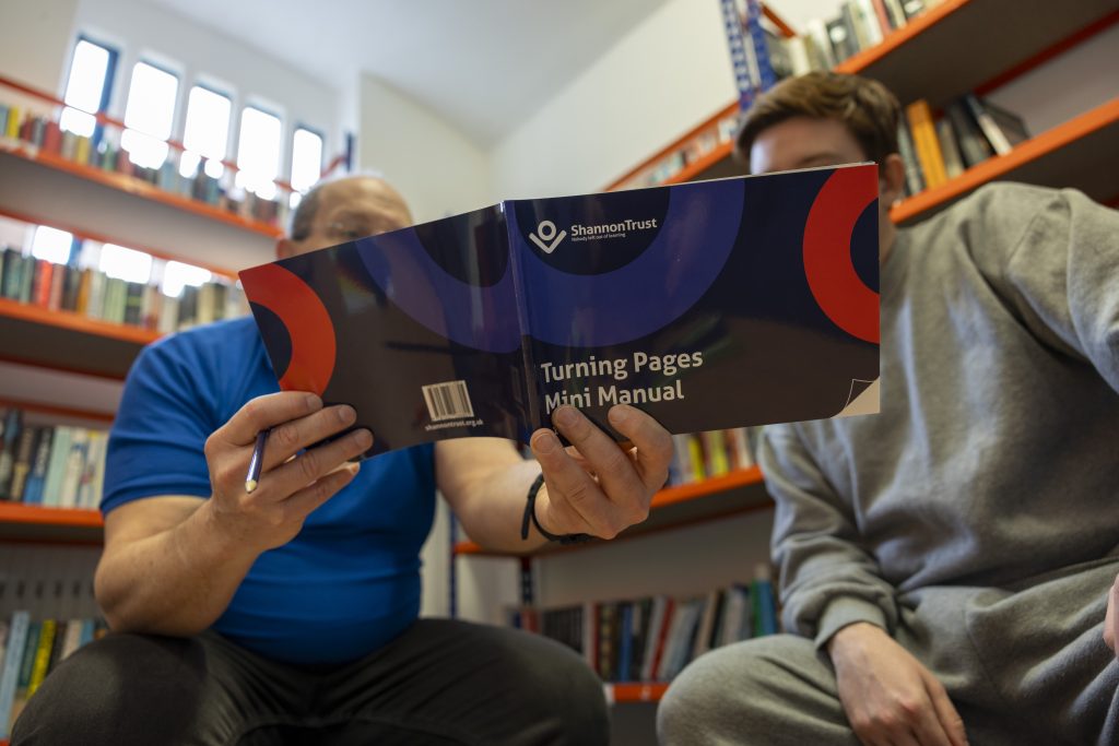 Two individuals sit facing each other in a library, focusing on a "Turning Pages Mini Manual" by Shannon Trust, which one person holds open. Together they are in the library of a prison, with bookshelves filled with books surrounding them.